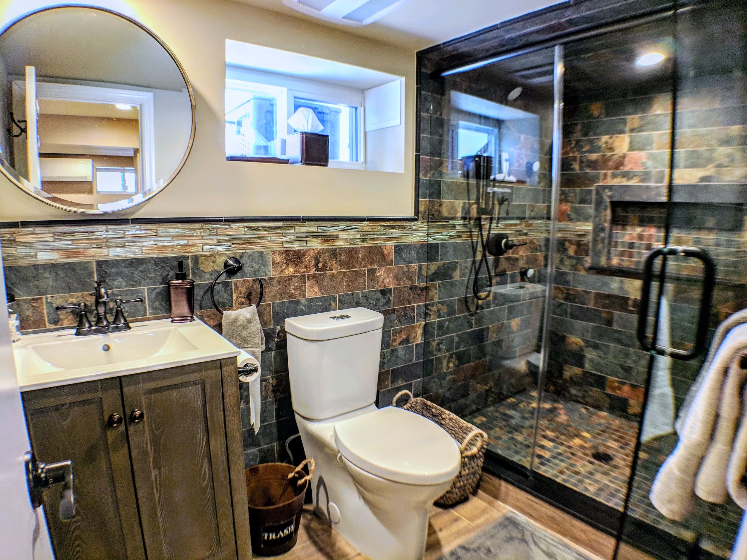 Basement bathroom with slate tile shower, round mirror, gray vanity